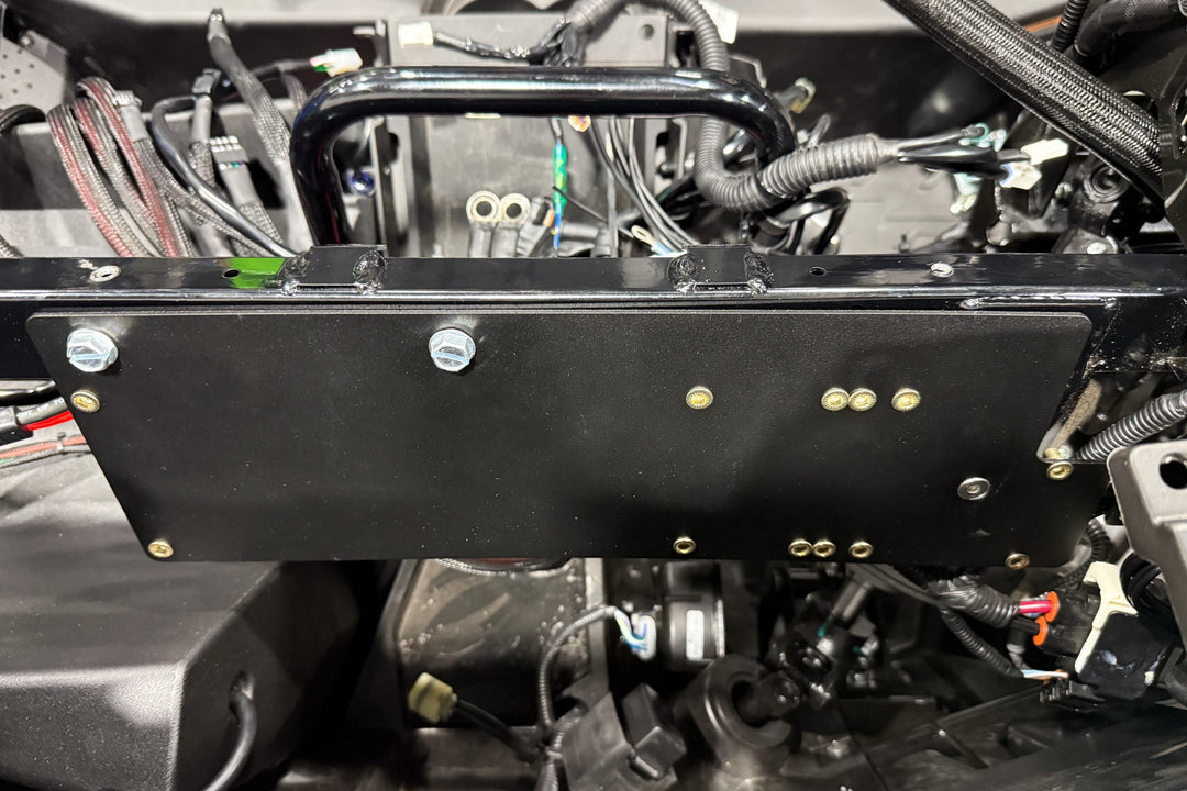 Black metal mounting plate installed on vehicle chassis with visible bolts and wiring harness
