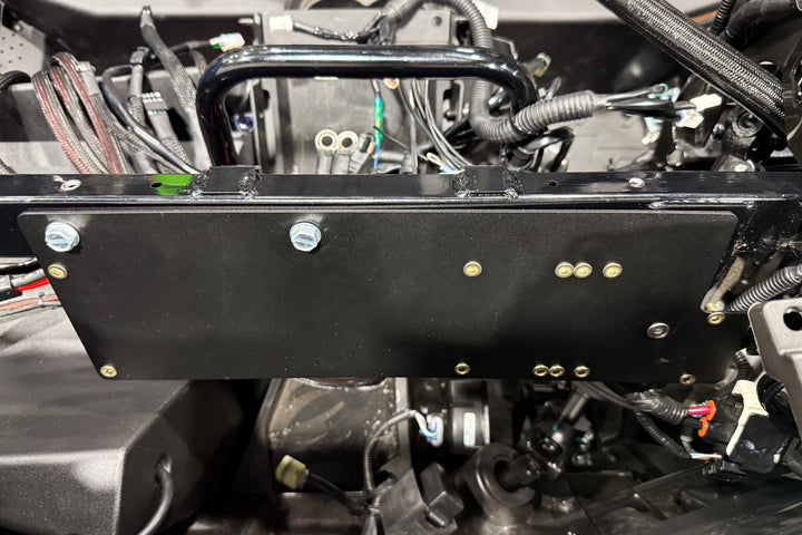 Black metal mounting plate installed on vehicle chassis with visible bolts and wiring harness